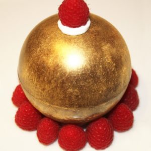 La Sphère d'Or, un dessert sphérique doré surmonté d'une seule framboise et entouré de huit framboises sur fond blanc, offre un délice visuel exquis.