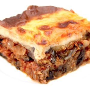 Une vue rapprochée d'une tranche de moussaka, mettant en valeur la cuisine grecque traditionnelle avec sa composition en couches de pommes de terre, d'aubergines, de viande hachée et de sauce béchamel crémeuse sur le dessus, sur une assiette blanche.