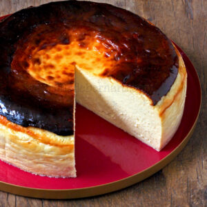 Un cheesecake basque brûlé entier dont une tranche a été retirée, révélant son intérieur crémeux, repose sur une assiette rouge sur une surface en bois.