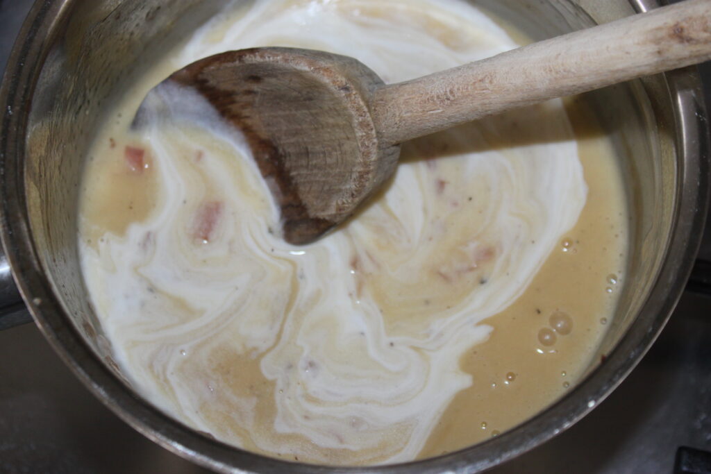 Une cuillère en bois remue un mélange crémeux dans une casserole, révélant des tourbillons de crème blanche mélangés à un liquide jaunâtre rappelant la sauce beurre blanc.