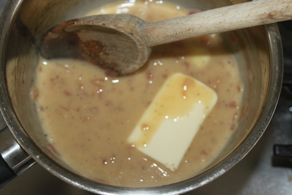 Un pot de sauce marron clair contient un bâton de beurre partiellement fondu et est remué avec une cuillère en bois, rappelant la préparation méticuleuse d'une sauce beurre blanc classique.