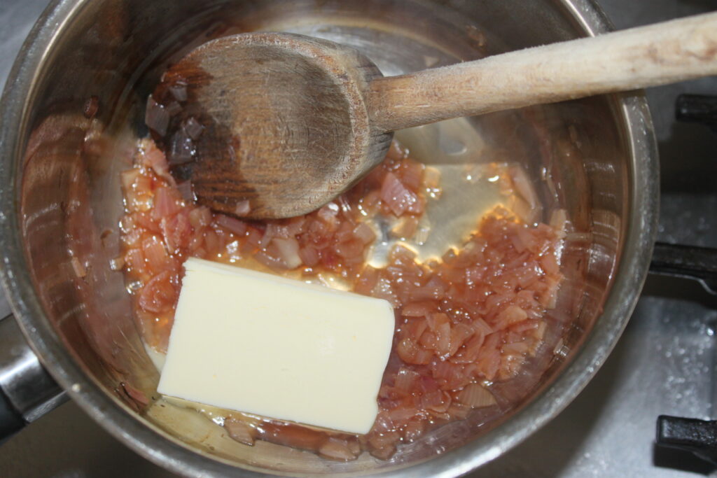 Une poêle en métal sur une cuisinière contient des oignons sautés, une cuillère en bois et une grosse tranche de beurre, se transformant en beurre blanc.