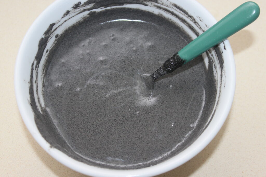 Bol blanc contenant un mélange liquide gris, rappelant le tahiné noir, avec une cuillère à manche vert partiellement enduite du mélange.