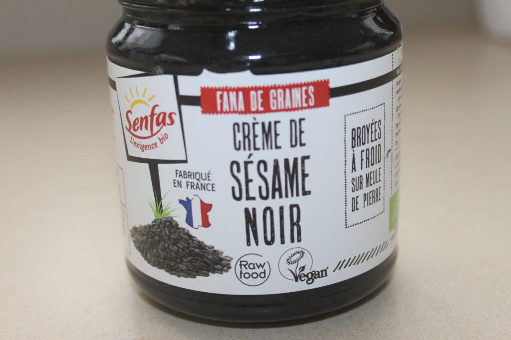 Un pot étiqueté « Crème de Sésame Noir », avec un texte indiquant qu'il est biologique, fabriqué en France, cru et végétalien. L'étiquette mentionne également qu'il est fabriqué à partir de graines de sésame noires moulues à froid. Cet exquis tahiné noir est parfait pour accompagner une sauce au beurre blanc sur des asperges fraîches.
