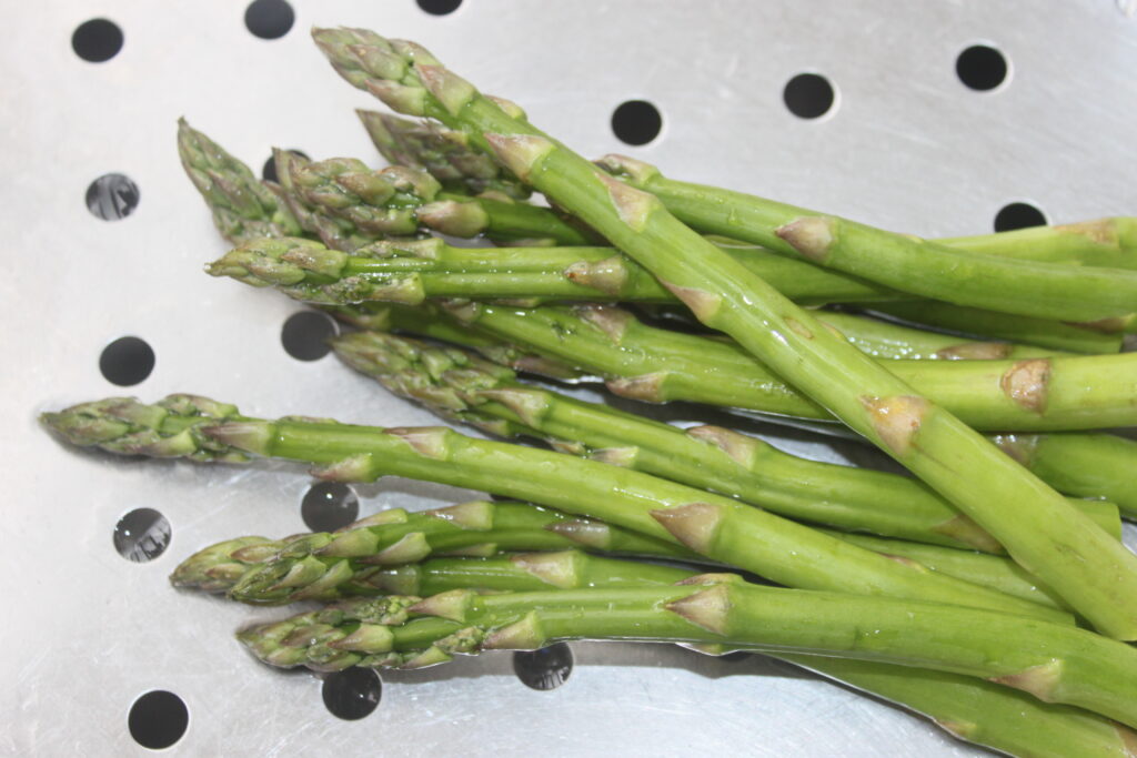 Plusieurs tiges d'asperges vertes fraîches reposent sur une surface métallique perforée, prêtes à être arrosées d'une riche sauce au beurre blanc.