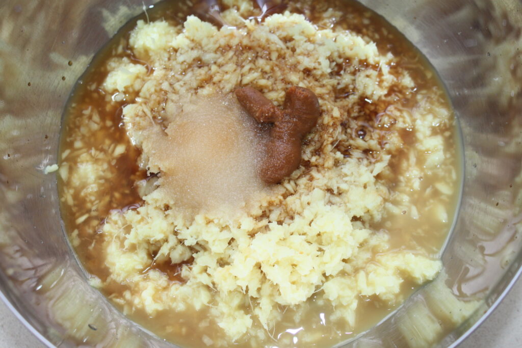 Un bol en métal contient un mélange d'ail émincé, de sauce soja, de pâte brune et de liquide. Les ingrédients, y compris éventuellement du gingembre pour plus de saveur, semblent être préparés pour la cuisson.