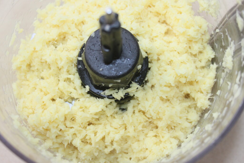 Gros plan du gingembre finement haché dans le bol d'un robot culinaire, avec la lame centrale visible, prêt à être transformé en une sauce savoureuse.