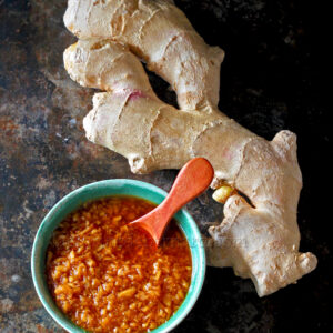 Un morceau de racine de gingembre frais se trouve à côté d'un bol de gingembre râpé dans une sauce brun rougeâtre, avec une cuillère en bois à l'intérieur du bol, le tout disposé sur une surface sombre.