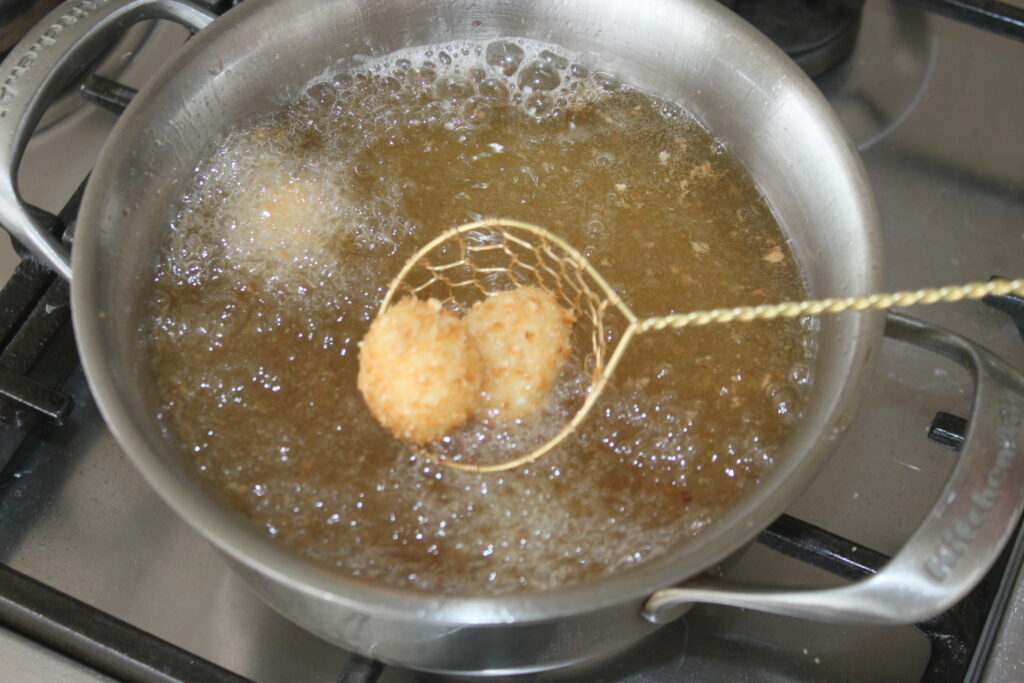 Une écumoire en métal soulève deux boules de mozza panées de l'huile chaude dans une poêle sur une cuisinière.