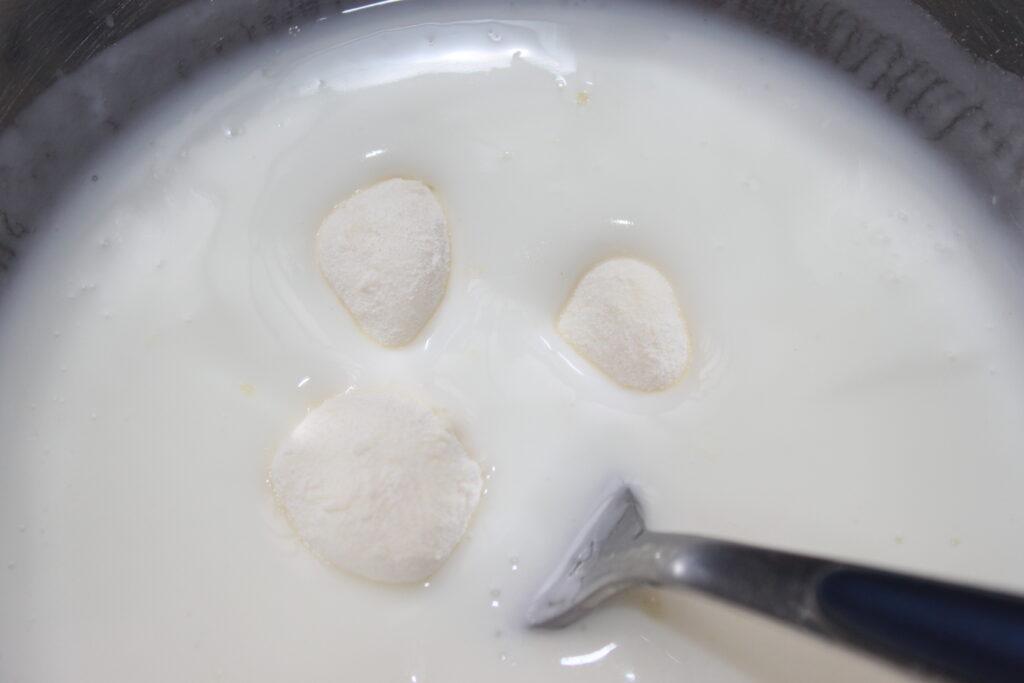 Trois morceaux de pâte flottent dans un bol de liquide blanc, ressemblant à des boules de mozza, avec une cuillère posée sur le côté droit.