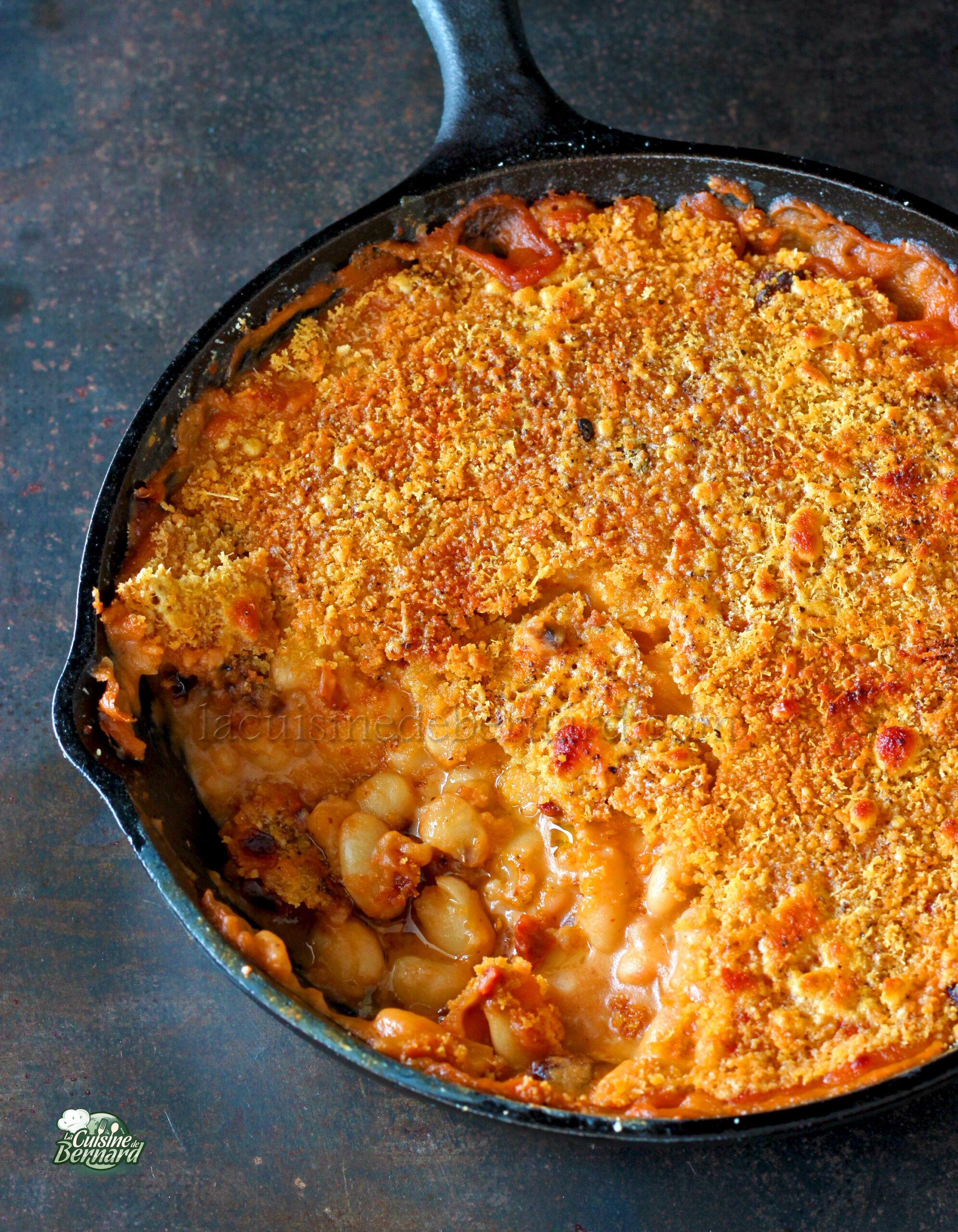 Une poêle en fonte remplie de macaronis au fromage cuits au four, garnie de chapelure dorée et croustillante et agrémentée de haricots blancs et de tomates séchées savoureuses.
