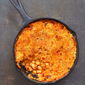 Une poêle en fonte remplie de haricots blancs à la sauce tomate, garnie d'une croûte de chapelure dorée croustillante et d'un soupçon de tomates séchées, avec une partie retirée pour révéler la garniture copieuse.