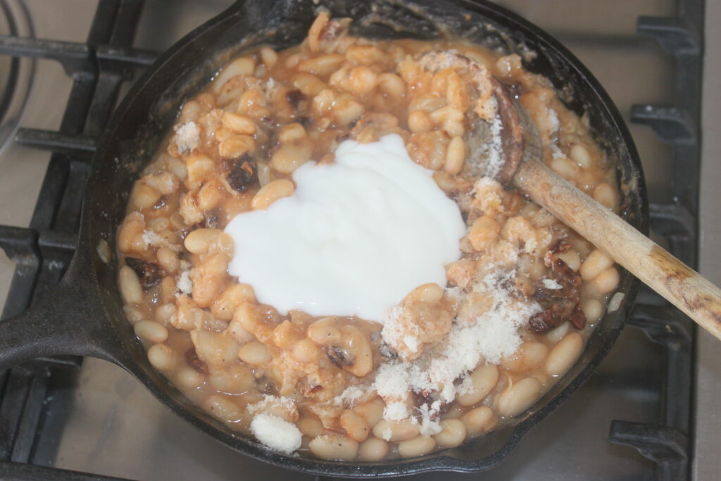 Une poêle avec des haricots blancs, de la sauce, des tomates séchées, une cuillerée de yaourt et du fromage râpé mélangés avec une cuillère en bois sur une cuisinière.