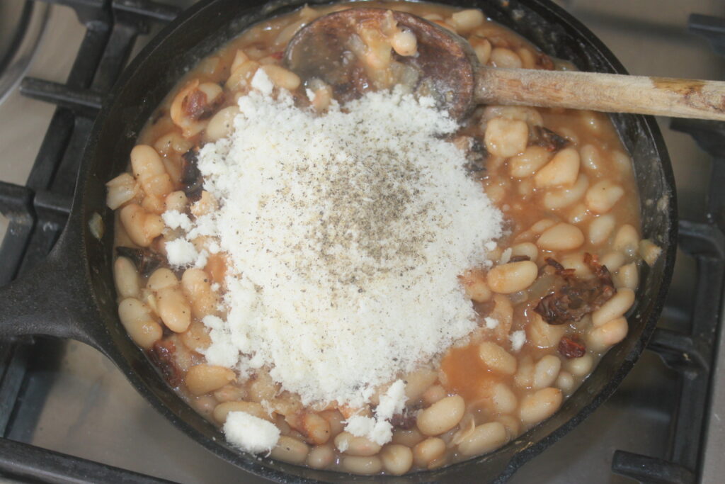 Une poêle en fonte sur une cuisinière contient des haricots blancs en sauce avec un gros tas de fromage râpé, de poivre noir et de tomates séchées sur le dessus, remués avec une cuillère en bois.