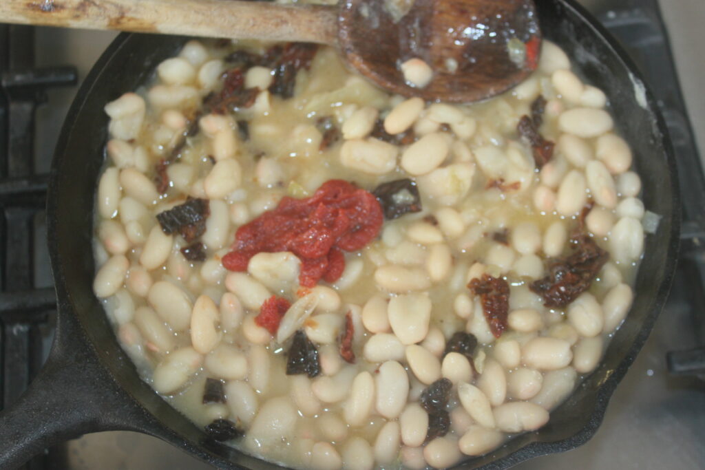 Une poêle en fonte de haricots blancs cuisant sur une cuisinière, avec une cuillère en bois, des morceaux visibles de concentré de tomates et des morceaux foncés de tomates séchées mélangés.