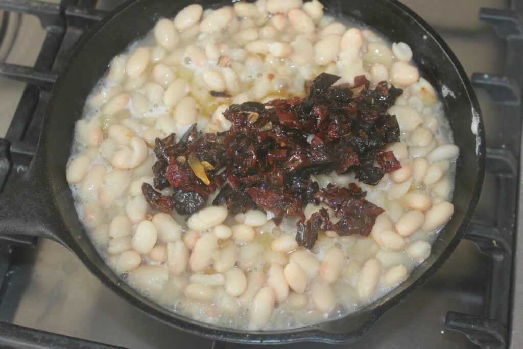 Une poêle en fonte sur une cuisinière contient des haricots blancs mijotant dans du liquide, garnis d'un tas de piments rouges séchés hachés et de tomates séchées éparpillées pour plus de profondeur de saveur.