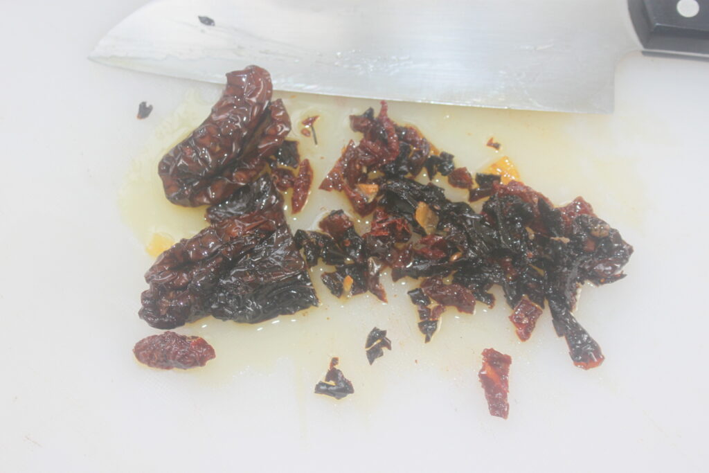 Tomates séchées hachées sur une surface blanche à côté d'un grand couteau de chef, prêtes à être ajoutées à des plats salés comme des haricots blancs.