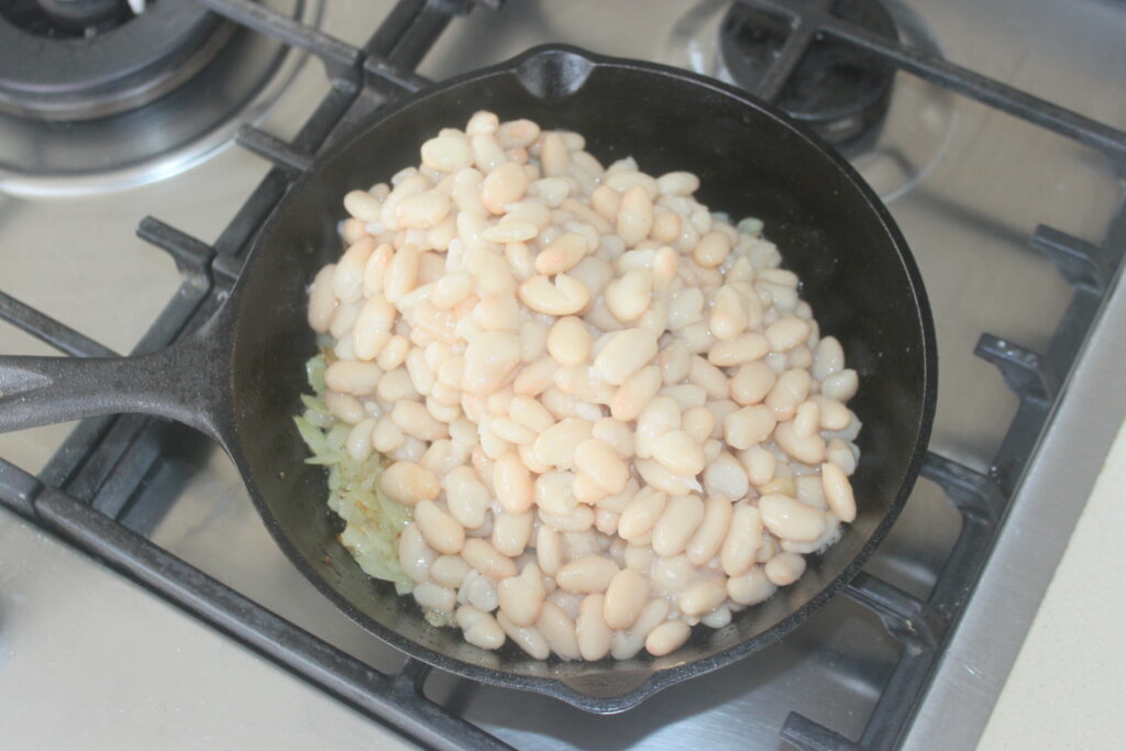Une poêle en fonte sur une cuisinière contient des haricots blancs empilés sur des oignons sautés, agrémentés de savoureuses tomates séchées.