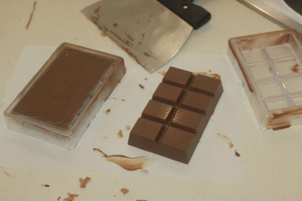 Une barre de chocolat retirée de son moule repose sur du papier sulfurisé, avec des moules à chocolat de luxe et un grattoir à proximité sur le comptoir.