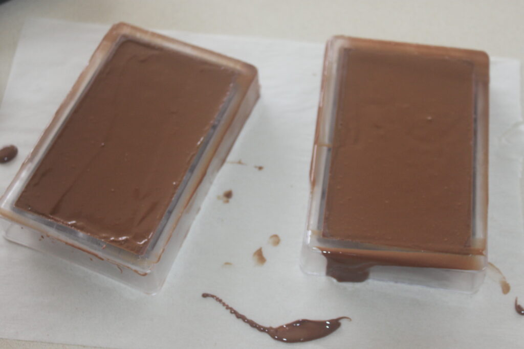Deux barres de chocolat de luxe dans des moules en plastique reposent sur une surface blanche, avec du chocolat fondu élégamment renversé autour d'elles.