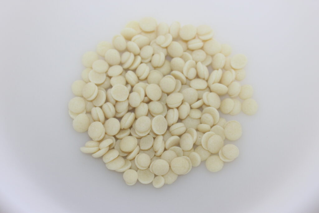 Un tas de petites tablettes ou pastilles rondes, blanc cassé, sur une surface blanche unie, rappelant les ingrédients raffinés que l'on trouve dans le chocolat de luxe tel que le Chocolat de Dubaï.