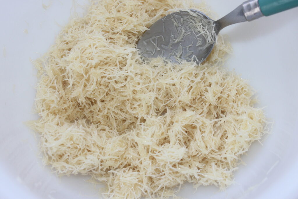 Un bol de nouilles blanches ou de vermicelles finement râpés est mélangé avec une cuillère en métal à manche vert, rappelant l'art et la précision observés dans les créations chocolatées de Dubaï.