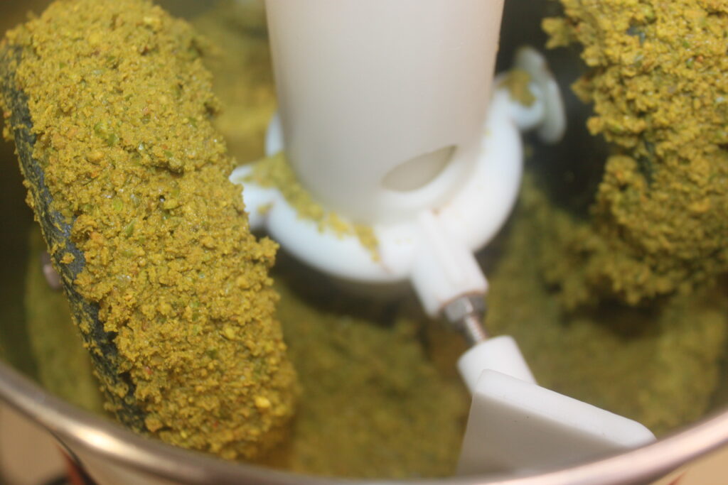 Gros plan d'un robot culinaire broyant un mélange verdâtre, peut être des noix ou des herbes, avec de la pâte collée aux lames et aux côtés parfait pour préparer les garnitures du Chocolat de Dubaï.