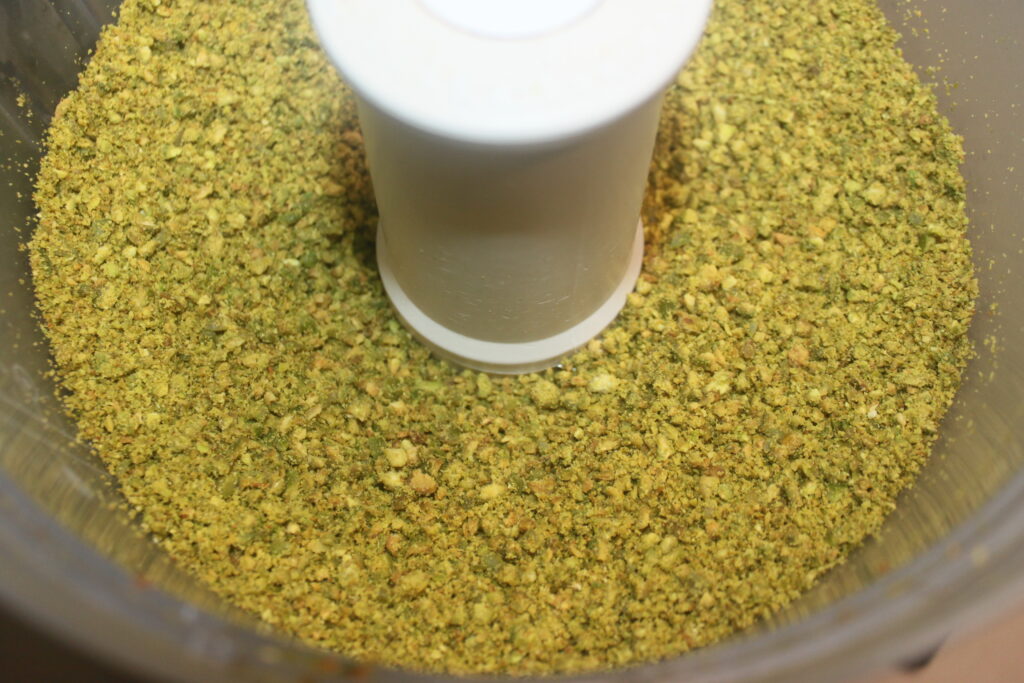 Pistaches moulues dans un bol de robot culinaire avec une lame blanche au centre, parfaites pour ajouter aux luxueux chocolats de Dubaï ou pour créer vos propres créations Chocolat de Dubaï.