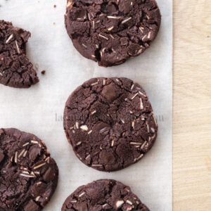 Quatre biscuits au chocolat entiers avec des morceaux de chocolat et des morceaux de noix sont disposés sur du papier sulfurisé Brouillon auto, avec une partie d'un biscuit cassé visible sur la gauche.