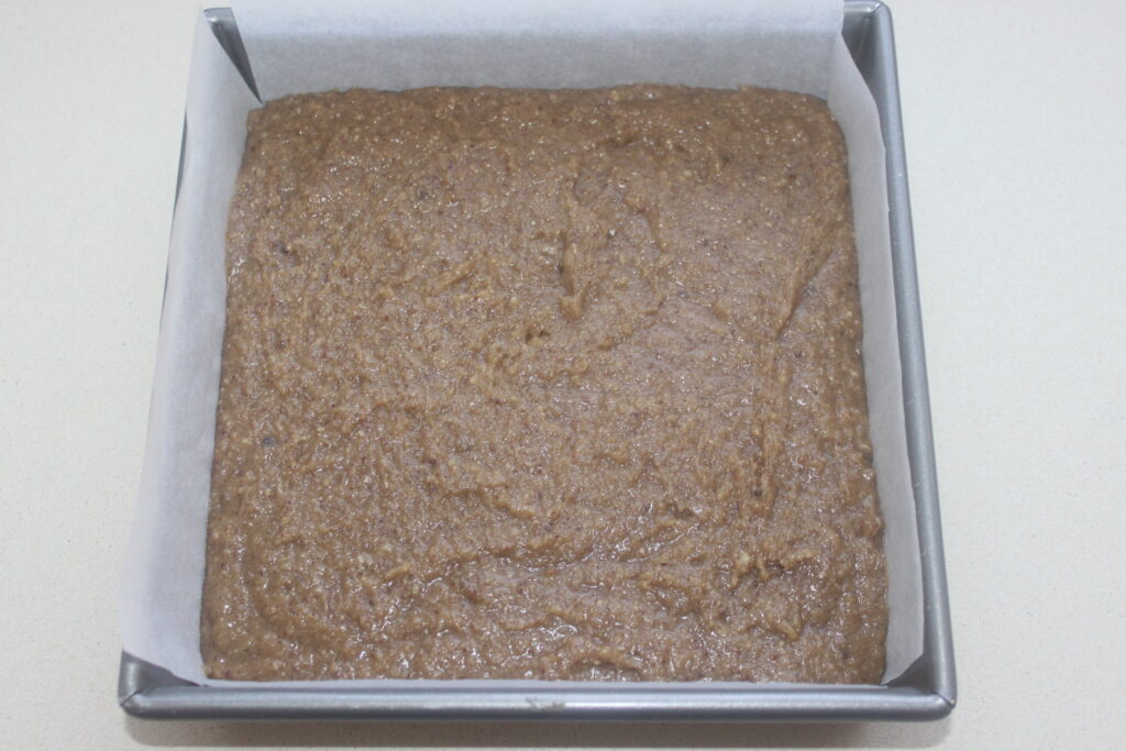 Moule à pâtisserie carré avec une pâte brune crue ressemblant à un riche financier, étalée uniformément et recouverte de papier sulfurisé.