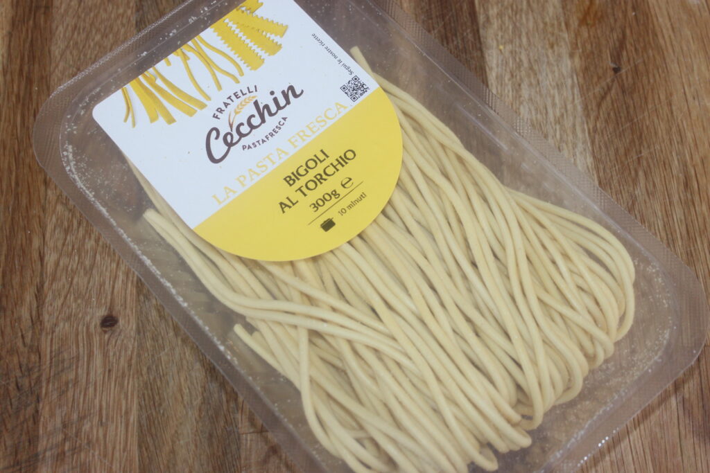 Un récipient en plastique de pâtes fraîches Fratelli Cochin Bigoli al Torchio de 300 g est posé sur un plan de travail en bois. Parfait pour accompagner votre salsa préférée.
