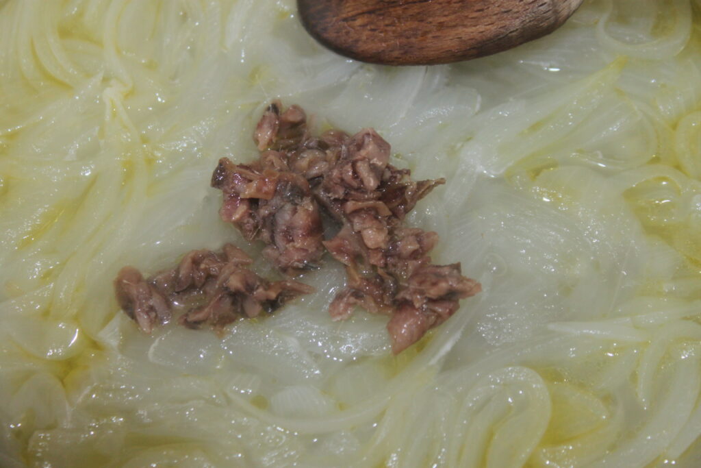 Des sardines hachées sur des oignons sautés à la poêle, rappelant la cuisine vénitienne classique, les bigoli en sauce. Une cuillère en bois est à portée de main, prête à donner vie à ce plat de pâtes.