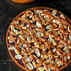 Une tarte ronde garnie de cacahuètes entières et arrosée de sauce au caramel repose élégamment sur une surface sombre.