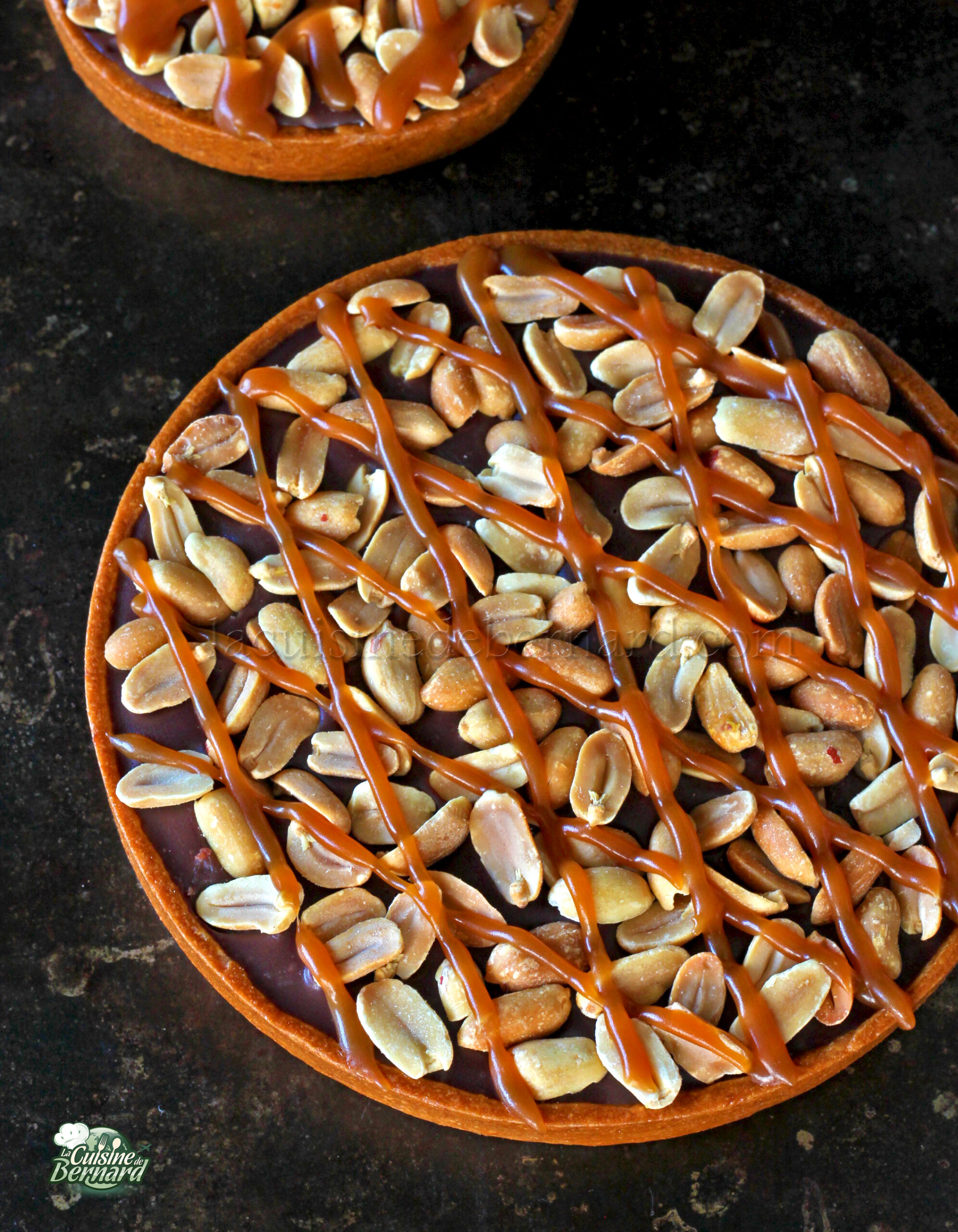 Une tarte ronde garnie de cacahuètes entières et arrosée de sauce au caramel repose élégamment sur une surface sombre.