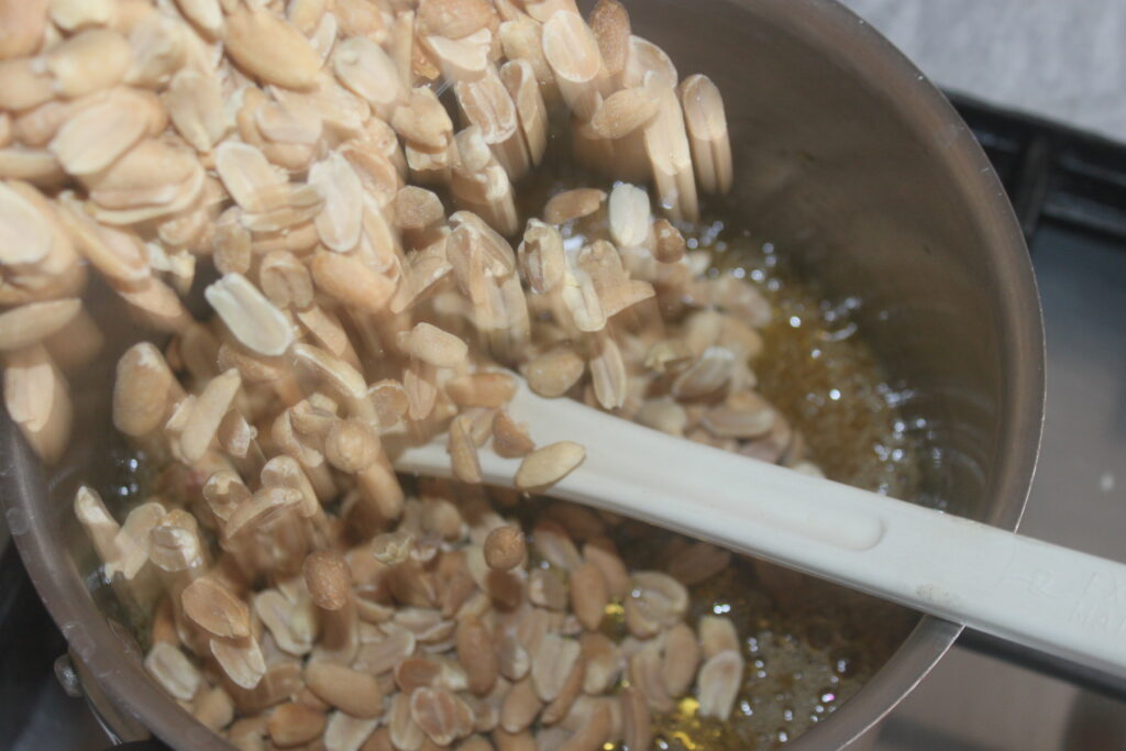 Des cacahuètes saupoudrées de chocolat tombent en cascade d'un récipient dans une casserole, avec une spatule posée à proximité, prête à remuer le délicieux mélange.