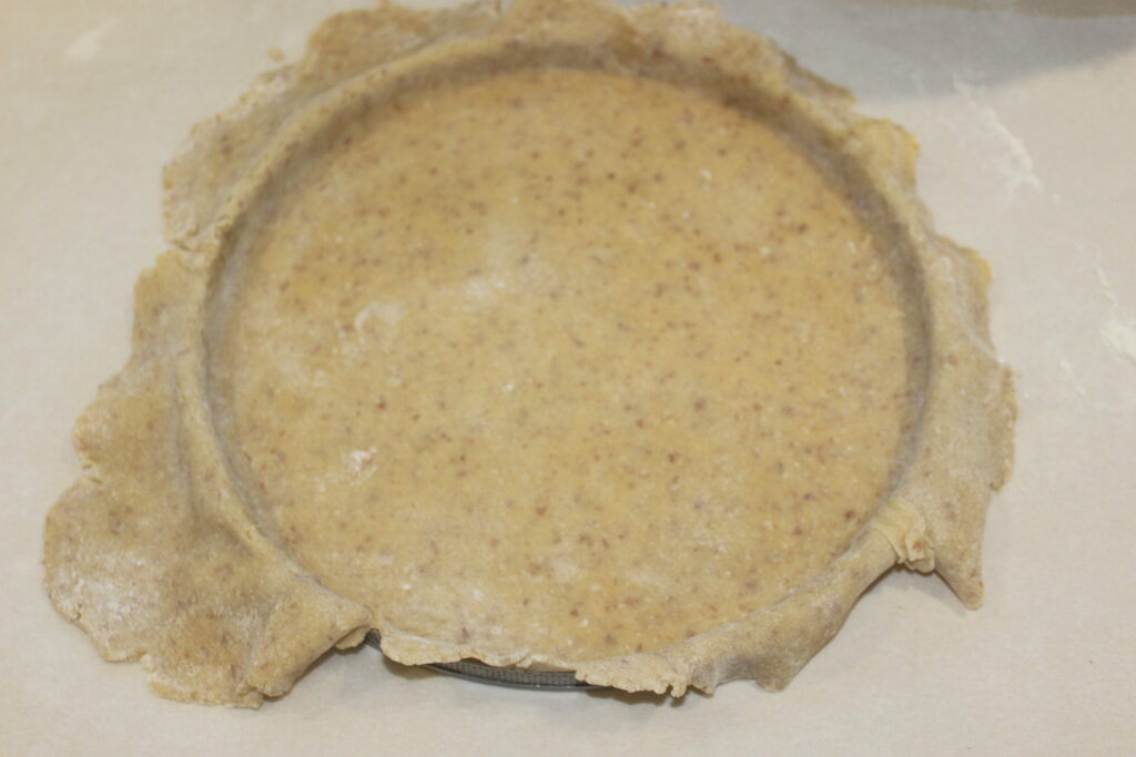 Une croûte de tarte rustique fabriquée à partir de pâte à grains entiers repose non cuite dans un moule rond sur une surface claire, prête à être remplie avec les ingrédients de votre choix.