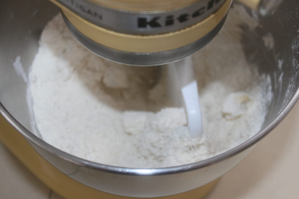 Un batteur sur socle équipé d'un crochet pétrisseur mélange efficacement la farine dans un bol en métal, prête à créer de délicieuses friandises infusées au chocolat ou aux cacahuètes.