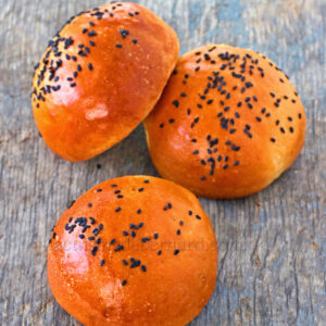 Trois petits pains moelleux dorés, garnis de graines de sésame noires, reposent élégamment sur une surface en bois.