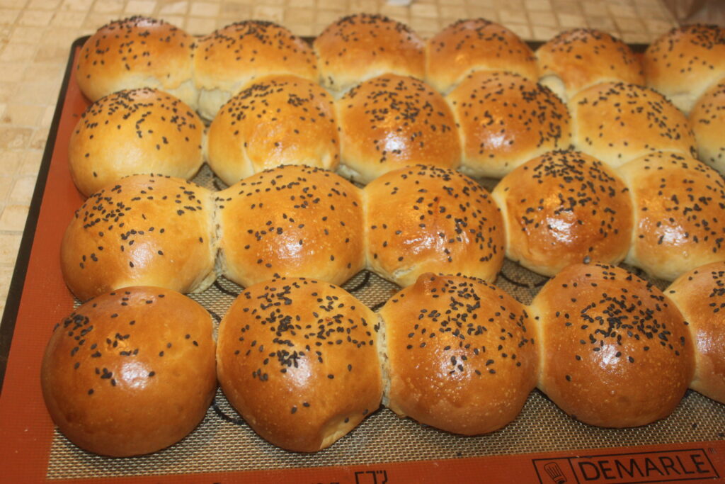 Un plateau de petits pains moelleux dorés, fraîchement cuits et uniformément espacés, garnis de graines de sésame noires.