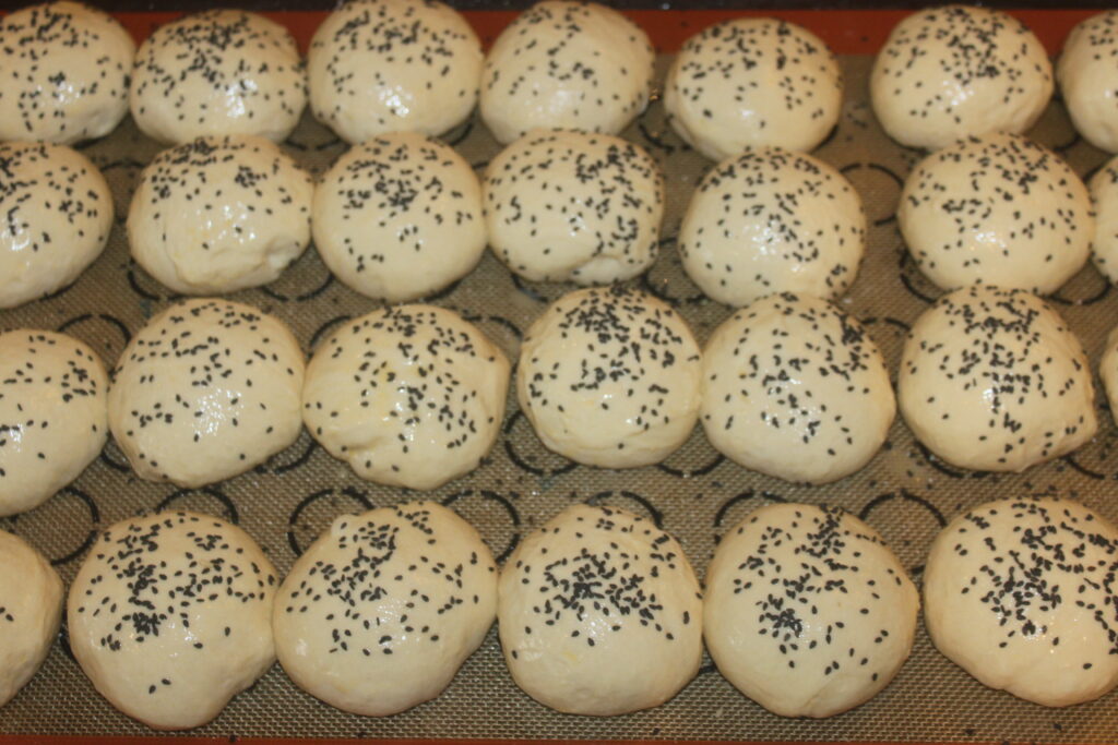 Des rangées de boules de pâte moelleuses, crues et généreusement garnies de graines de sésame noires, reposent sur un tapis de cuisson.