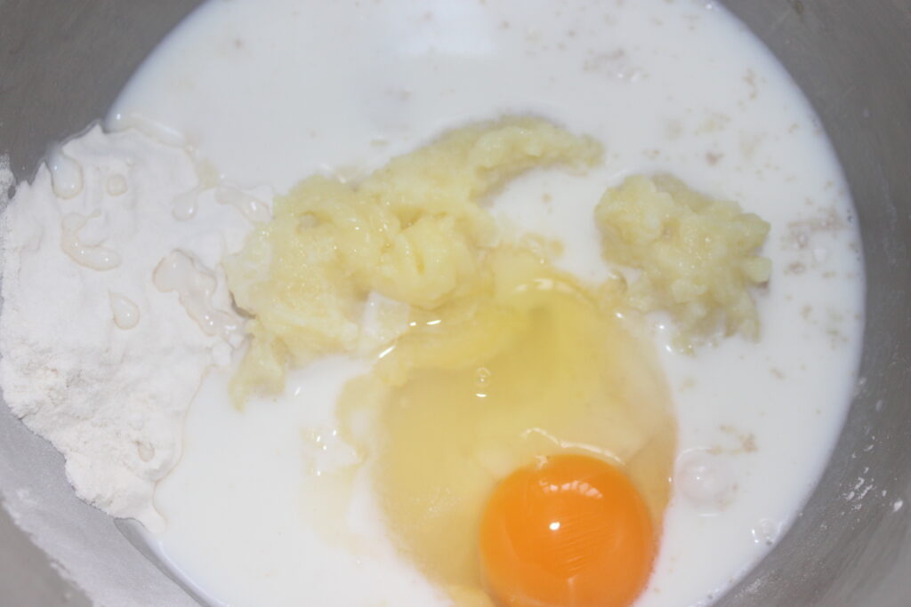 Un bol à mélanger contient de la farine, des bananes écrasées, du lait et un œuf cassé, créant la base parfaite pour des petits pains moelleux et moelleux.