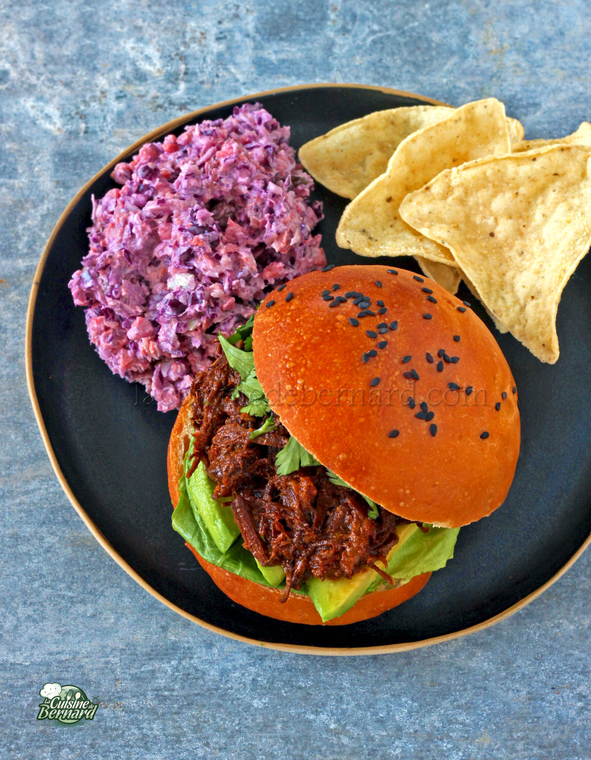 Sandwich au porc effiloché avec laitue et avocat sur un pain au sésame, servi avec salade de chou violette et chips de tortilla sur une assiette noire.