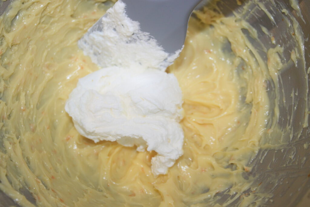 Un bol à mélanger contient une pâte jaune crémeuse mélangée à une cuillerée de crème blanche à l'aide d'une spatule, rappelant la riche garniture des tartelettes à la crème d'orange.