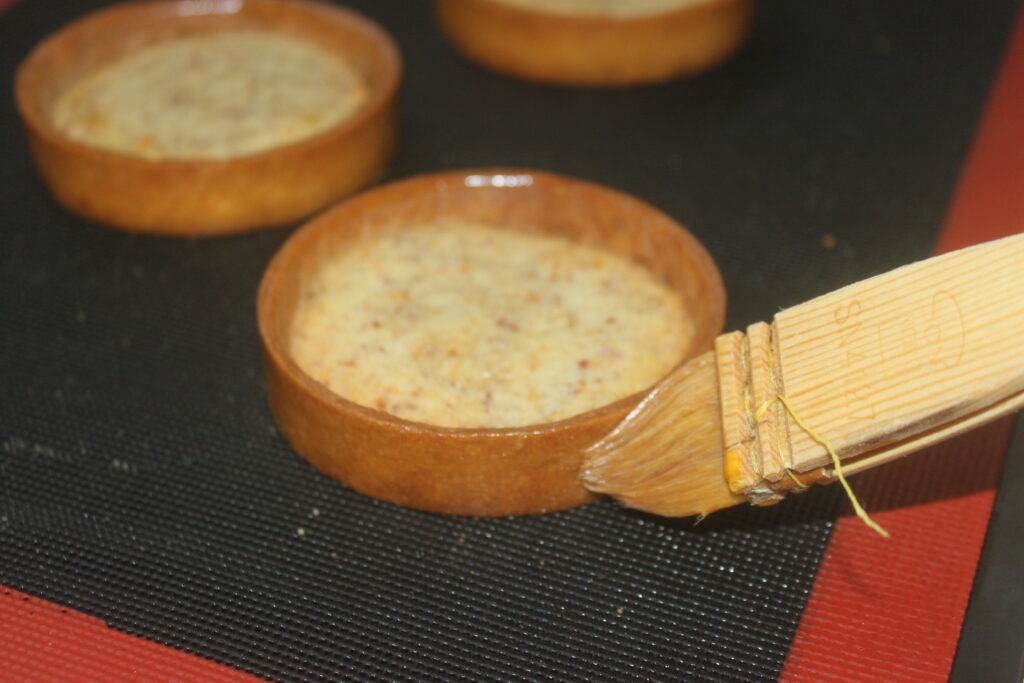 De délicates coques de pâtisserie, badigeonnées de dorure à l'œuf à l'aide d'un pinceau en bois sur une plaque à pâtisserie, sont prêtes à devenir d'exquises tartelettes crème d'orange fourrées de crème d'orange crémeuse pour un délicieux dessert.