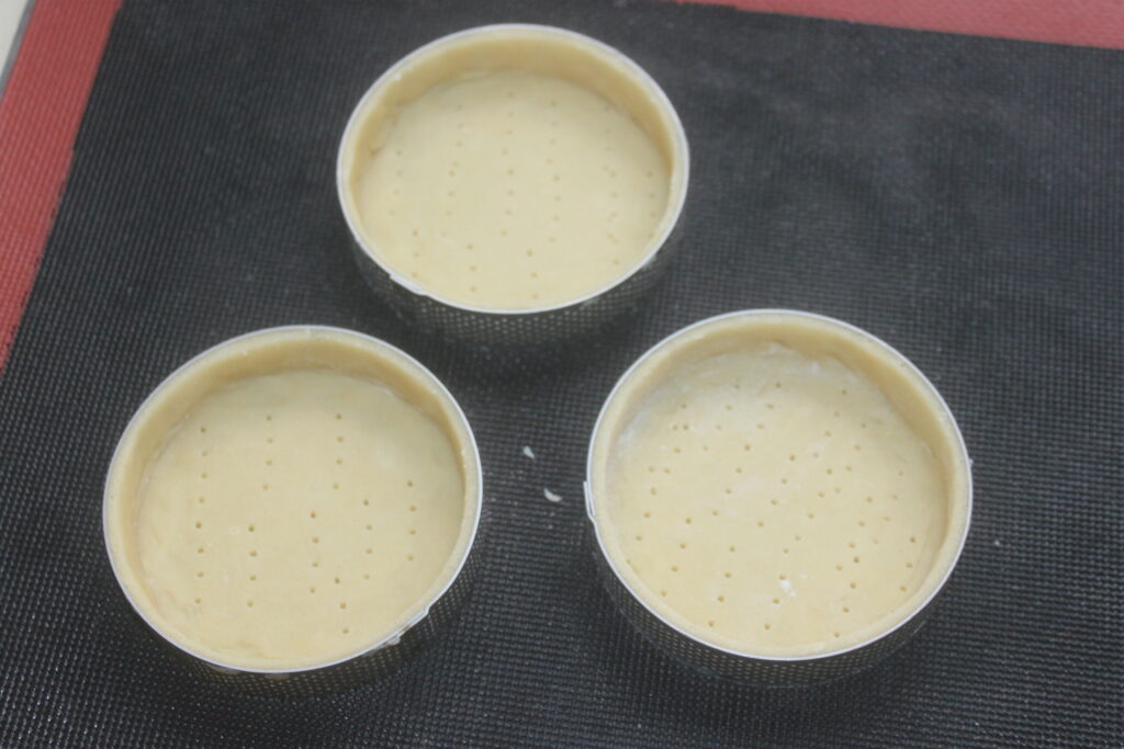 Trois tartelettes crème d'orange crues aux fonds piqués sont disposées sur un plan noir, prêtes à être garnies.