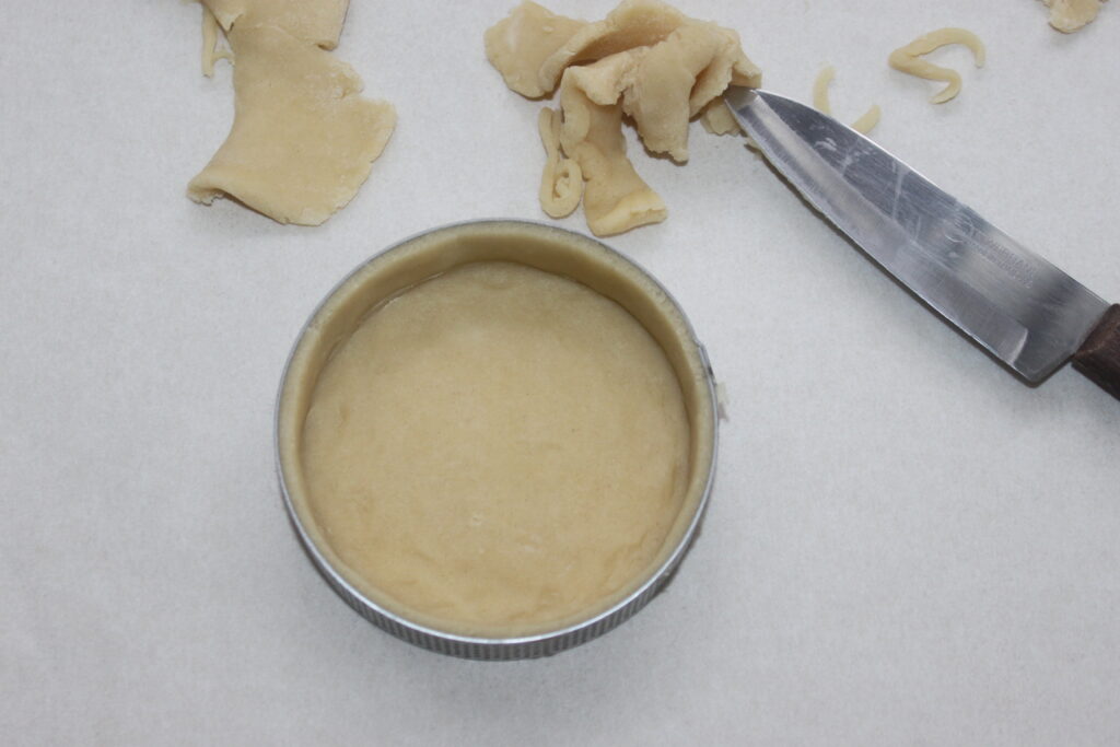 La pâte des Tartelettes à la crème d'orange est placée dans un cercle en métal sur du papier sulfurisé, avec de la pâte supplémentaire et un couteau à proximité, prête à être transformée en délicieuses friandises.