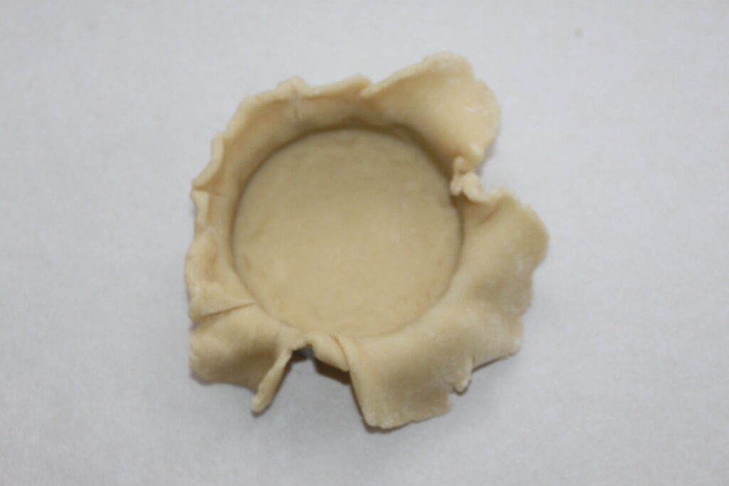 Une croûte de pâtisserie non cuite aux bords irréguliers et surplombants est posée dans un plat de cuisson sur une surface claire, constituant la base idéale pour les Tartelettes crème d'orange.