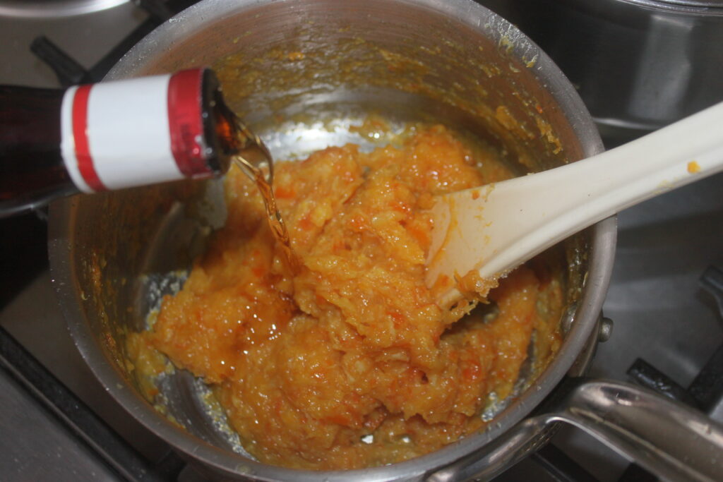 Le liquide coule d'une bouteille dans une casserole, se mélangeant aux légumes oranges remués par une cuillère blanche, évoquant la création de Tartelettes crème d'orange.