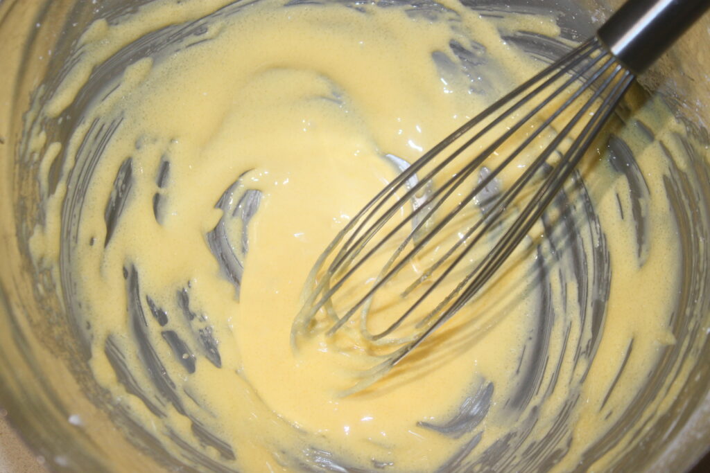 Un fouet en métal mélange élégamment la pâte jaune pâle dans un bol en verre, destinée à devenir des Tartelettes crème d'orange, des desserts exquis infusés de l'essence vibrante de la crème d'orange.