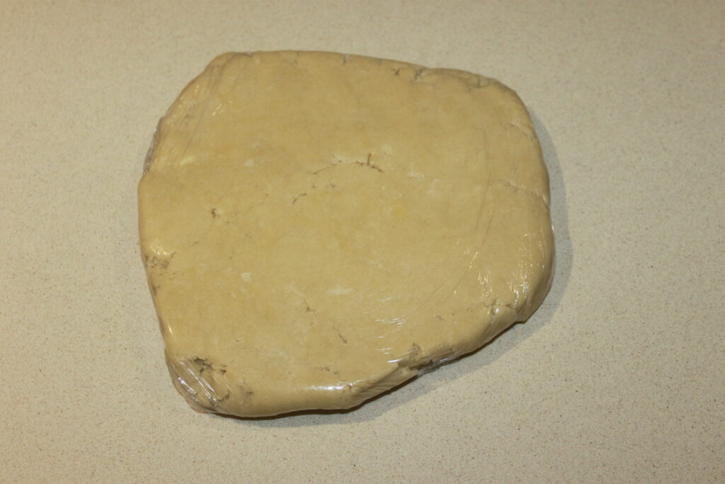 Une pâte ronde et aplatie enveloppée dans du plastique transparent sur une surface beige est prête à devenir des Tartelettes crème d'orange.